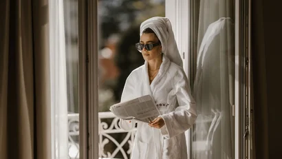 Woman in bathrobe with towel on head reading newspaper on balcony
