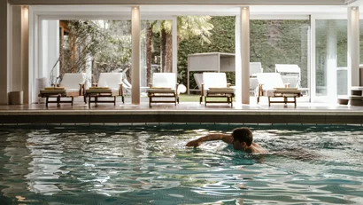 Person swimming in indoor pool with lounge chairs and garden view