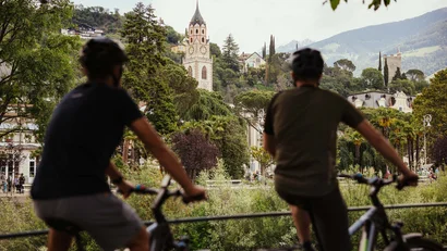 Two cyclists overlooking a town with a church tower and castle in the mountains