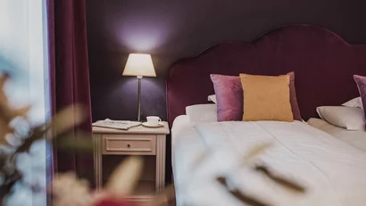 Lit bedside table with lamp, newspaper, and cup next to bed with purple headboard