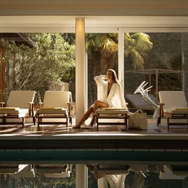 Woman in bathrobe sitting on lounge chair next to indoor pool with plants in background