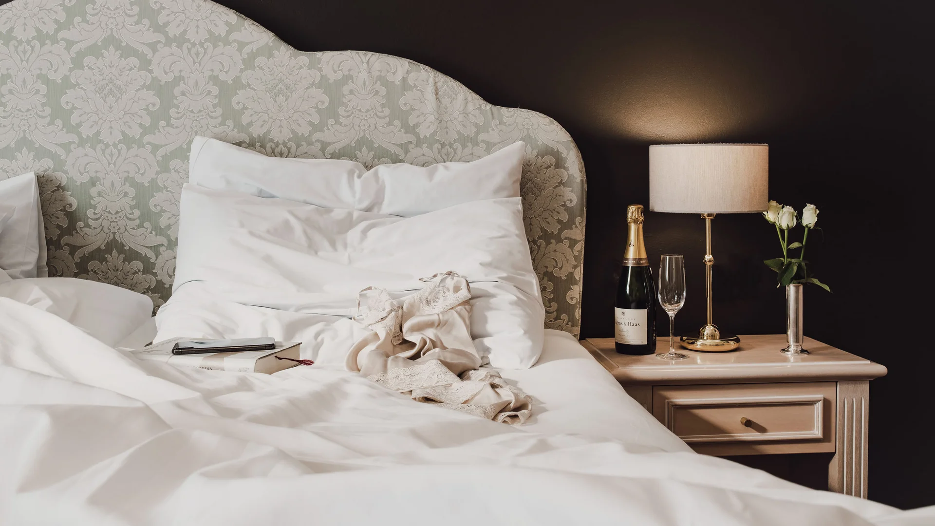 Gemachtes Bett mit Musterkopfteil, Champagnerflasche, Glas und Lampe auf Nachttisch
